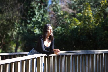 Esmer bir kadın, genç ve güzel günlük giysiler içinde tahta bir köprünün parmaklıklarına yaslanmış. Kadın farklı vücut duruşları sergiliyor. Arka planda yeşil bitki örtüsü