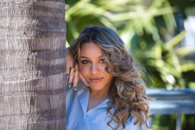 Sarışın, genç ve güzel bir kadın. Günlük giysiler içinde palmiye ağacının gövdesine yaslanmış. Kadın mutlu ve kameraya bakıyor.