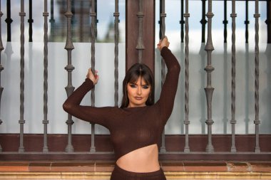 Dudaklarından ve elmacık kemiklerinden estetik ameliyat geçirmiş genç bir kadın pencerenin ızgarasını tutuyor. Kadın fotoğraf için kameraya bakıyor.