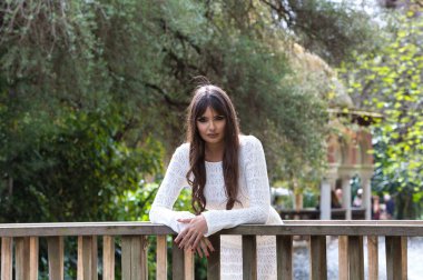 Beyaz örgü elbiseli, genç ve güzel esmer kadın, parktaki bir köprünün ahşap korkuluklarına yaslanarak farklı ifadeler sergiliyor. Arka planda ağaçlar ve bitki örtüsü