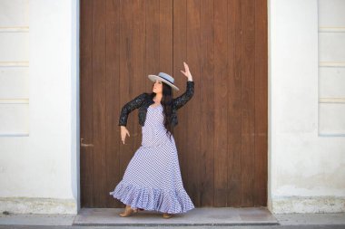 Latin bir kadın, Endülüs 'ün tipik bir sokağındaki ahşap bir kapının önünde dans eden genç ve güzel bir flamenko. Kadın, siyah benekli, şapkalı ve ceketli beyaz bir elbise giyiyor.