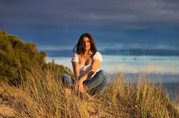 Genç ve güzel Latin kadın, esmer, sazlıkların arasında beyaz bir kum tepesinde oturuyor. Kadın mutlu ve gülümsüyor. Arka planda deniz ve bulutlu mavi gökyüzü.