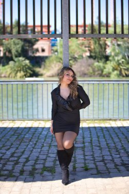 Siyah giyinmiş genç sarışın kadın mutlu ve metal posterlere yaslanıyor. Arka planda nehir Seville, Endülüs şehrinden geçerken. Kadın güneşli günün tadını çıkarıyor.