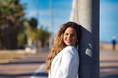 Genç ve güzel sarışın bir kadının portresi sokak lambasına yaslanmış ve kameraya bakıyor, kadın mutlu ve gülümsüyor ve beyaz bir ceket giyiyor.