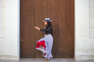 Latin bir kadın, Endülüs 'ün tipik bir sokağındaki ahşap bir kapının önünde dans eden genç ve güzel bir flamenko. Kadın, siyah benekli, şapkalı ve ceketli beyaz bir elbise giyiyor.