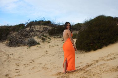 Zarif turuncu elbiseli genç ve güzel esmer kadın İspanya 'nın Endülüs sahilindeki güzel kumulların arasında yürüyor. Kadın farklı duruşlar sergiliyor.