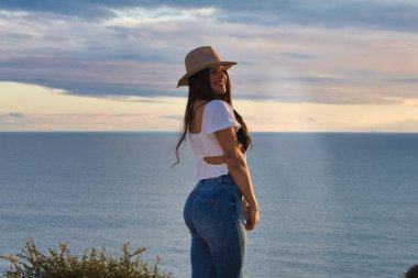 Latin kadın, esmer, genç ve güzel ince beyaz kumdan bir uçurumda. O, kot pantolon ve hasır şapka giydi. Kadın gülümser ve gün batımında altın saatinde kameraya bakar.