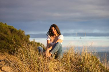 Genç ve güzel Latin kadın, esmer, sazlıkların arasında beyaz bir kum tepesinde oturuyor. Kadın mutlu ve gülümsüyor. Arka planda deniz ve bulutlu mavi gökyüzü.