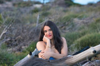 Genç, güzel, mavi iç çamaşırlı bir Latin kadın gün batımının altın saatinde bir ormanda duruyor. Kadın mutlu ve gülümsüyor ve kuru bir kütüğe yaslanıyor.