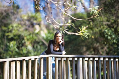 Esmer bir kadın, genç ve güzel günlük giysiler içinde tahta bir köprünün parmaklıklarına yaslanmış. Kadın farklı vücut duruşları sergiliyor. Arka planda yeşil bitki örtüsü