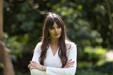 İspanyol esmer, genç ve güzel bir kadın, kollarını kavuşturuyor ve güçlü ve havalı bir tavırla ileriye bakıyor. Kadın ormanda, ağaçların arasında.