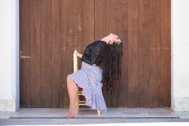 Latin kadın, genç ve güzel, dans eden flamenko, eski tahta bir sandalyede oturuyor ve kafasını geriye atıyor. Kadın, siyah benekli ve siyah ceketli, beyaz, dalgalı bir elbise giyiyor.