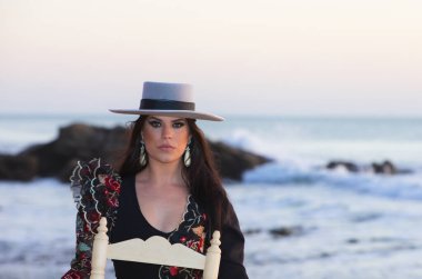 Güzel esmer İspanyol kadın tipik bir kostüm ve geniş kenarlı şapkayla flamenko yapıyor arka planda eski bir tahta sandalyede oturuyor Atlantik Okyanusu ve kayaları.