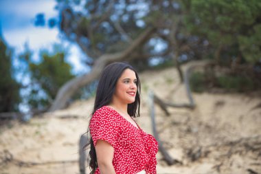 Kumsalın oradaki kumullarda çıplak ayaklı genç, esmer kadın. Kadın gülümsüyor ve mutlu. Kadın beyaz pantolon ve kırmızı gömlek giyiyor.