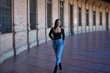 Esmer bir kadın, kot pantolon giymiş genç ve güzel ve siyah bir elbise giymiş Seville, Endülüs 'teki İspanya' nın ünlü meydanında yürüyor. Kadınlar anıtın tadını çıkarıyor.