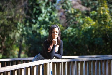 Esmer bir kadın, genç ve güzel günlük giysiler içinde tahta bir köprünün parmaklıklarına yaslanmış. Kadın farklı vücut duruşları sergiliyor. Arka planda yeşil bitki örtüsü