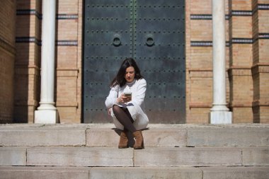 Genç ve güzel Latin esmer kadın merdivenlerde oturmuş cep telefonuna bakıyor. Kadın dikkatle cep telefonu ekranına bakıyor.