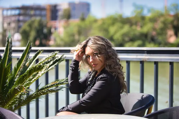 Siyah giyinmiş güzel sarışın kadın Seville şehrinin nehir kıyısındaki terasta oturuyor. Arka planda şehir ve bitki örtüsü var. Kadın farklı ifadeler kullanıyor.