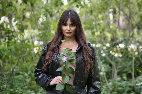 İspanyol kadın portresi, uzun saçlı esmer kadın ve ormanda bir seans için göğsünün arasında çiçekle poz veren tek siyah ceket. Arka planda bitki örtüsü ve ağaçlar