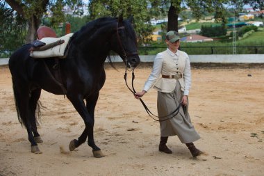 Genç ve güzel bir kadın Endülüs, İspanya 'da siyah safkan bir İspanyol atının dizginlerini tutuyor ve çekiyor. Kadın tipik bir atlı kadın kostümü giymiş.