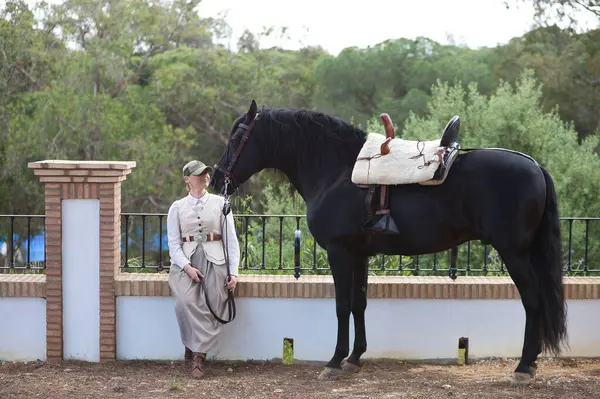 İspanya, Endülüs 'te siyah safkan İspanyol atının yanında bir çam ağacının gölgesinde oturan güzel genç kadın. Kadın tipik bir atlı kadın kostümü giymiş.