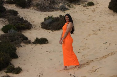 Zarif turuncu elbiseli genç ve güzel esmer kadın İspanya 'nın Endülüs sahilindeki güzel kumulların arasında yürüyor. Kadın farklı duruşlar sergiliyor.