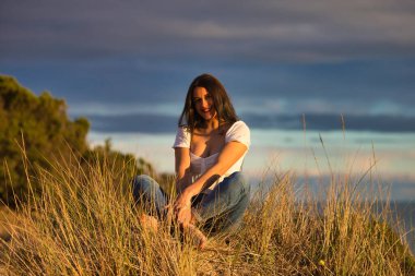 Genç ve güzel Latin kadın, esmer, sazlıkların arasında beyaz bir kum tepesinde oturuyor. Kadın mutlu ve gülümsüyor. Arka planda deniz ve bulutlu mavi gökyüzü.