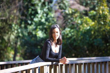 Esmer bir kadın, genç ve güzel günlük giysiler içinde tahta bir köprünün parmaklıklarına yaslanmış. Kadın farklı vücut duruşları sergiliyor. Arka planda yeşil bitki örtüsü