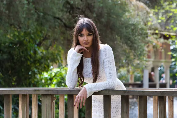 Beyaz örgü elbiseli, genç ve güzel esmer kadın, parktaki bir köprünün ahşap korkuluklarına yaslanarak farklı ifadeler sergiliyor. Arka planda ağaçlar ve bitki örtüsü