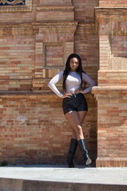 Genç ve güzel bir Afrikalı kadın, modern kıyafetler giymiş, elleri kalçasında, İspanya 'nın Seville meydanındaki kare kırmızı tuğla bir sütunun yanında dinleniyor. Seyahat ve tatil konsepti