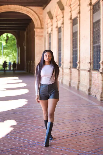 Deri şort, siyah çizme ve bej bluz giyen genç ve güzel Afrikalı kadın tatilde ve İspanya 'nın ünlü meydanında Seville' de yürüyor. Seyahat ve tatil konsepti