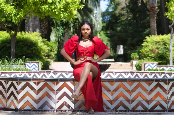 Afrikalı bir kadın, uzun kahverengi saçlı, genç ve güzel, zarif kırmızı bir elbise giymiş, Seville 'de altın bir fıskiyenin kenarında çapraz bacaklı oturuyor. Kadın farklı pozlar veriyor.