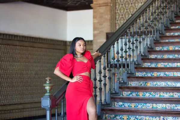 Afrikalı bir kadın, uzun kahverengi saçlı, genç ve güzel, zarif kırmızı bir parti elbisesi içinde Endülüs, Seville 'deki İspanyol meydanının merdivenlerinde.