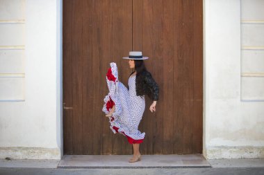 Latin bir kadın, Endülüs 'ün tipik bir sokağındaki ahşap bir kapının önünde dans eden genç ve güzel bir flamenko. Kadın, siyah benekli, şapkalı ve ceketli beyaz bir elbise giyiyor.