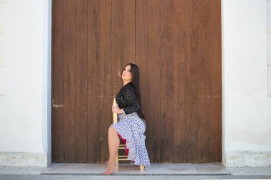 Latin kadın, genç ve güzel, dans eden flamenko, eski bir tahta sandalyede oturuyor. Kadın, siyah benekli, kısa siyah ceketli, beyaz, dalgalı bir elbise giyiyor.