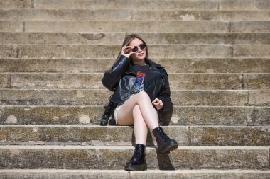 Güneş gözlüğü takan ve rockçı gibi giyinmiş güzel sarışın bir kadın merdivenlerin merdivenlerinde bacak bacak üstüne atmış oturuyor. Kadın huzur içinde dinleniyor.