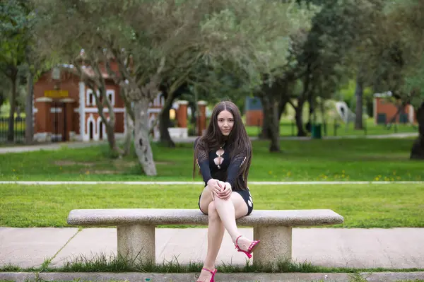Latin bir kadın, siyah ve pembe topuklu giyen genç ve güzel bir esmer bir bankta bağdaş kurmuş oturuyor. Kadın farklı ifadeler ve jestler yapıyor. Arka planda zeytin ağaçları