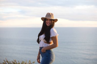 Latin kadın, esmer, genç ve güzel ince beyaz kumdan bir uçurumda. O, kot pantolon ve hasır şapka giydi. Kadın gülümser ve gün batımında altın saatinde kameraya bakar.