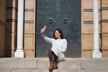 Genç ve güzel Latin esmer kadın merdivenlerde oturmuş selfie çekiyor. Kadın dikkatle mobil ekrana bakıyor ve fotoğraf için gülümsüyor.