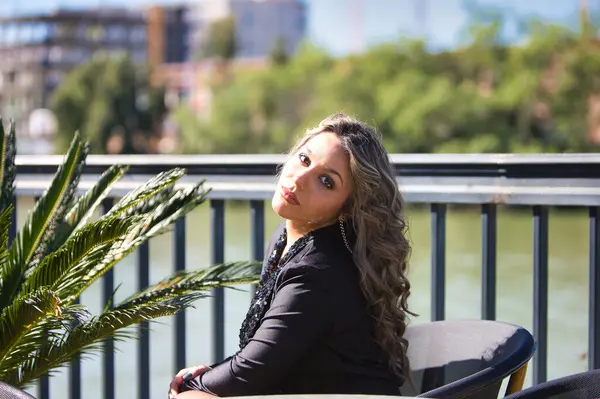 Siyah giyinmiş güzel sarışın kadın Seville şehrinin nehir kıyısındaki terasta oturuyor. Arka planda şehir ve bitki örtüsü var. Kadın farklı ifadeler kullanıyor.
