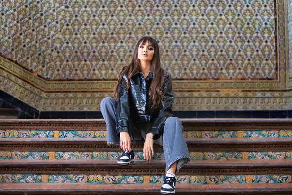 İspanya, Seville ve Endülüs 'ün ünlü meydanının merdivenlerinde siyah deri ceketli ve kot pantolonlu güzel esmer kadın oturuyor. Arkaplanda Arap tarzı fayanslar