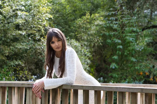 Beyaz örgü elbiseli, genç ve güzel esmer kadın, parktaki bir köprünün ahşap korkuluklarına yaslanarak farklı ifadeler sergiliyor. Arka planda ağaçlar ve bitki örtüsü