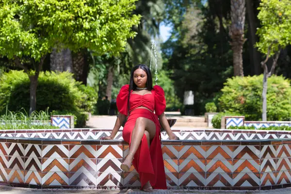 Afrikalı bir kadın, uzun kahverengi saçlı, genç ve güzel, zarif kırmızı bir elbise giymiş, Seville 'de altın bir fıskiyenin kenarında çapraz bacaklı oturuyor. Kadın farklı pozlar veriyor.