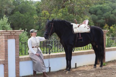 İspanya, Endülüs 'te siyah safkan İspanyol atının yanında bir çam ağacının gölgesinde oturan güzel genç kadın. Kadın tipik bir atlı kadın kostümü giymiş.