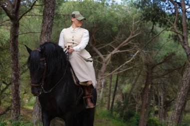 Genç ve güzel bir kadın çam ormanında safkan bir İspanyol siyah atına biniyor. Kadın tipik bir atlı kadın kostümü giyiyor. Arka planda yemyeşil bitki örtüsü