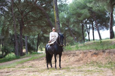 Genç ve güzel bir kadın çam ormanında safkan bir İspanyol siyah atına biniyor. Kadın tipik bir atlı kadın kostümü giyiyor. Arka planda yemyeşil bitki örtüsü