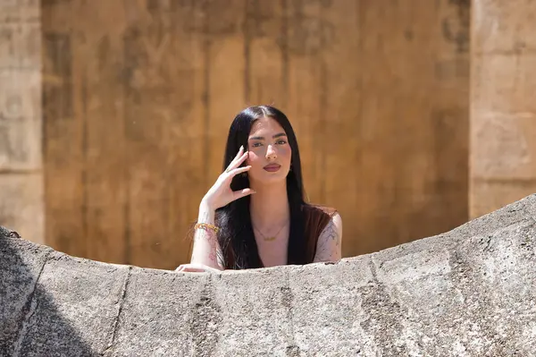 Uzun saçlı, genç, güzel esmer bir kadın yanağını elinde dinlendiriyor ve düşünceli ve bitkin görünüyor. Kadın farklı yönlere bakıyor. Arka planda kahverengi bir duvar var