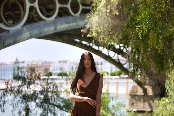 Uzun saçlı, kahverengi elbiseli ve siyah çizmeli çekici bir kadın Sevilla 'daki Triana Köprüsü' nün yanındaki nehir kıyısında duruyor.
