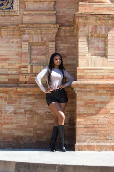 Genç ve güzel bir Afrikalı kadın, modern kıyafetler giymiş, elleri kalçasında, İspanya 'nın Seville meydanındaki kare kırmızı tuğla bir sütunun yanında dinleniyor. Seyahat ve tatil konsepti