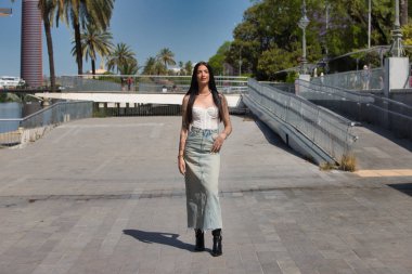 Kollarında dövmeleri olan, sıradan kıyafetler giyen, güneşli bir günde büyük bir şehir caddesinde poz veren genç ve çekici bir Latin kadın. Kadın makyaj yapmış.